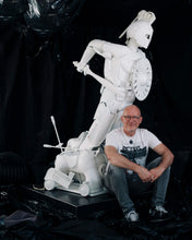 Load image into Gallery viewer, Artist Scoobafish seated beside Plastic Apocalypse – Trojan Warrior, showing the scale of the white sculpture made from reclaimed objects.