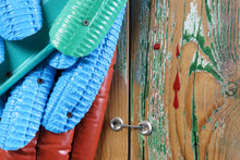Load image into Gallery viewer, Close-up of blue, green and red plastic feathers and a metal fastener on the weathered wooden boards of Proud Parrot by Scoobafish.
